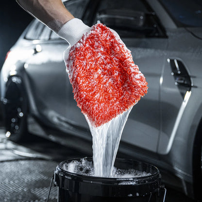 Een hand houdt een zeepachtige microvezel washand boven een emmer voor een grijze auto, zodat autolak veilig wordt gereinigd met groot absorberend vermogen.