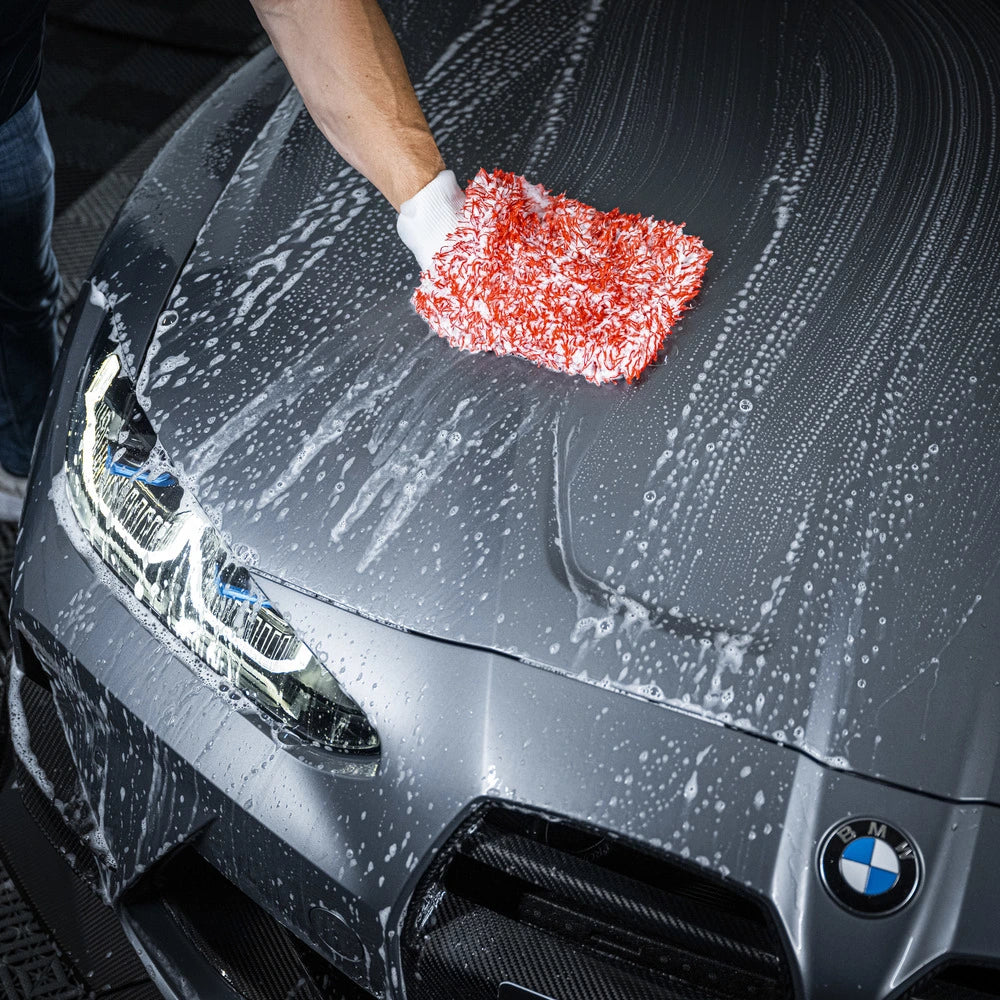 Een persoon gebruikt een microvezel washandschoen met groot absorberend vermogen om de motorkap van een grijze BMW auto autolak veilig te reinigen.