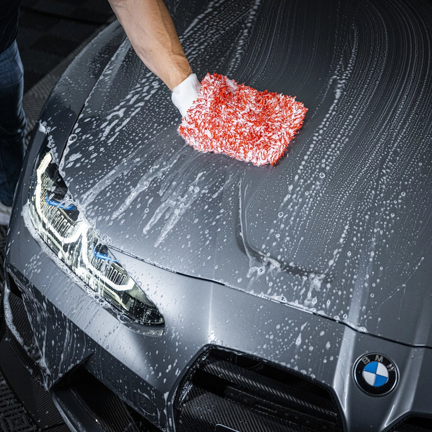 Een persoon gebruikt een microvezel washandschoen met groot absorberend vermogen om de motorkap van een grijze BMW auto autolak veilig te reinigen.