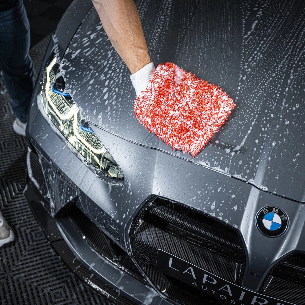Een persoon gebruikt autoshampoo en een zeepachtige rood-witte handschoen om een grijze BMW motorkap schoon te maken, met water en zeep op het oppervlak.