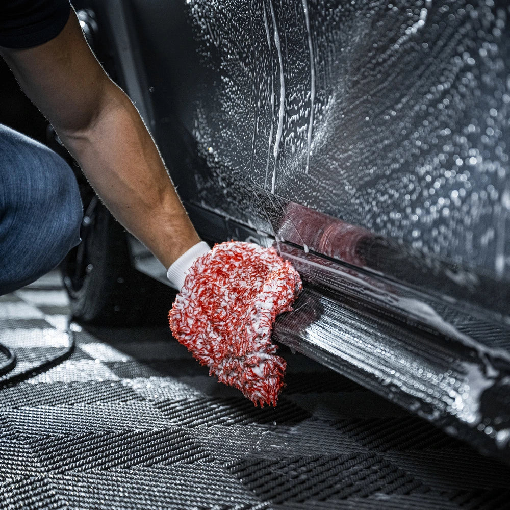 A person uses a white glove and red-and-white cleaning mitt from the Starter Kit | 21-delig to autowassen the lower side of a car door, covered in soapy water—a close-up showcases the careful autopoetsen process.