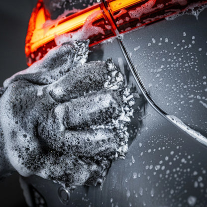 Een gehandschoende hand met een microvezel washandschoen zeept de achterkant van een zilveren auto in, waardoor de autolak veilig blijft; boven is rood achterlicht zichtbaar.