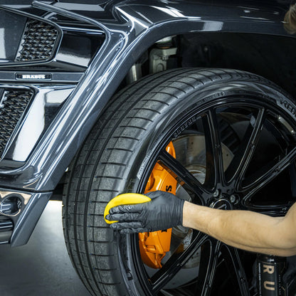 Een persoon met een zwarte handschoen brengt rubber verzorging aan op een autoband met een gele spons bij een zwart wiel en oranje remklauw.