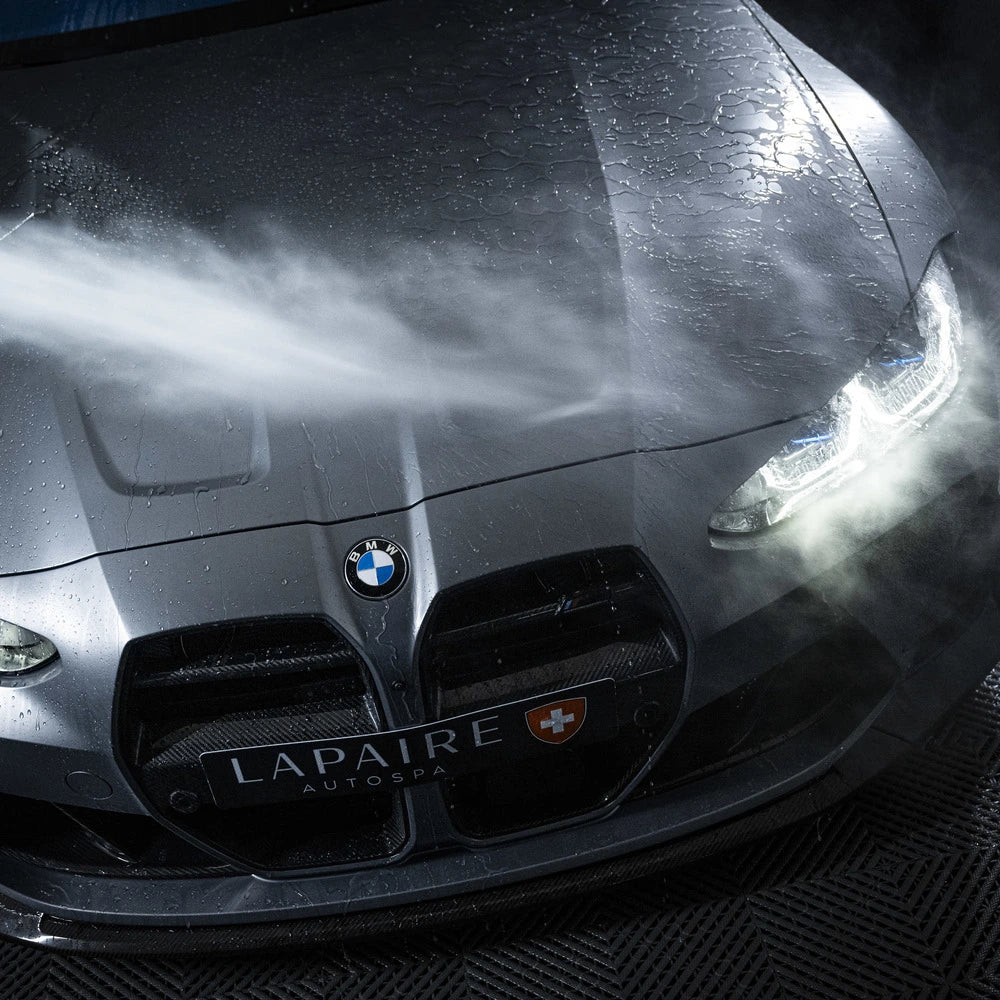 A close-up of a wet black BMW hood being sprayed with water, headlights on and droplets visible—showcasing professional autopoetsen with the Starter Kit | 21-delig from LAPAIRE AUTOSPA.