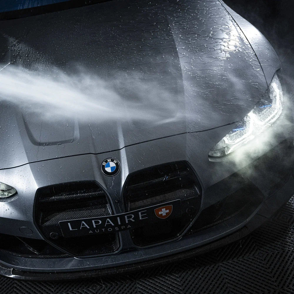 A close-up of a black BMW’s front being sprayed with water during a wash, showing water beading after applying 1+1 GRATIS Nano Wet Coat. The headlights are illuminated and the LAPAIRE AUTOSPA license plate is visible.