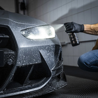 Een persoon met een zwarte handschoen spuit insectenverwijderaar op de koplamp van een grijze auto in een garage, klaar voor autoreiniging.