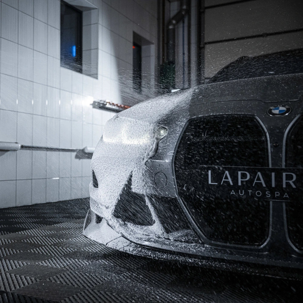 Een BMW auto bedekt met een dikke laag sneeuwschuim wordt gewassen in een overdekte spa, waarbij water op de voorkant spuit.
