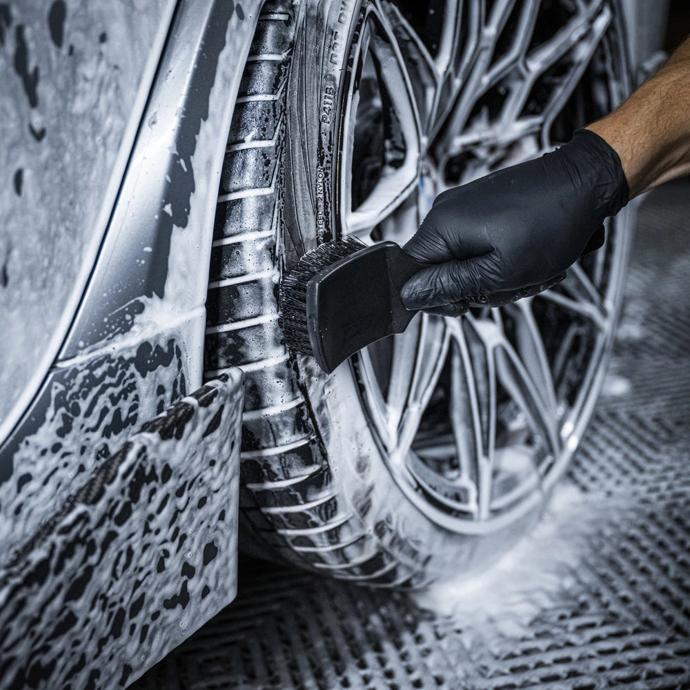 Wearing a black glove, a person uses a brush from the Starter Kit XXL | 25-delig to scrub a car tire, creating soap suds on the tire, wheel, and nearby surface.