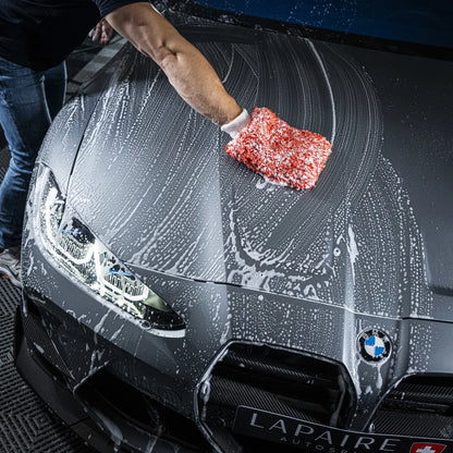 Using the Starter Kit XXL | 25-delig, a person washes the hood of a grey BMW with a red and white sponge, creating soap suds. The car’s headlights and part of the LAPAIRE license plate are visible in the scene.