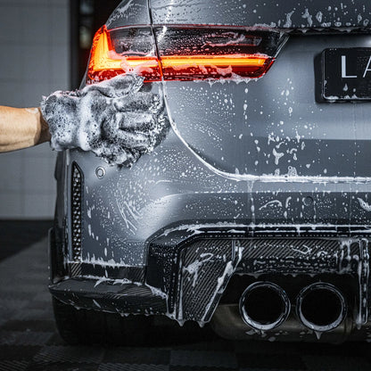 Een hand van een persoon gebruikt een microvezel washandschoen om de achterkant van een zilveren auto met zeepsop te wassen; het achterlicht is verlicht.
