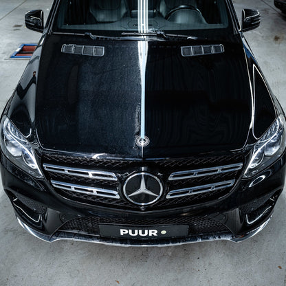 A black Mercedes-Benz in a garage shows a bold dirt line down the hood, highlighting the dramatic effect of the Ceramic Sealant Package. The PUUR license plate underscores the contrast between protected and unprotected sides.