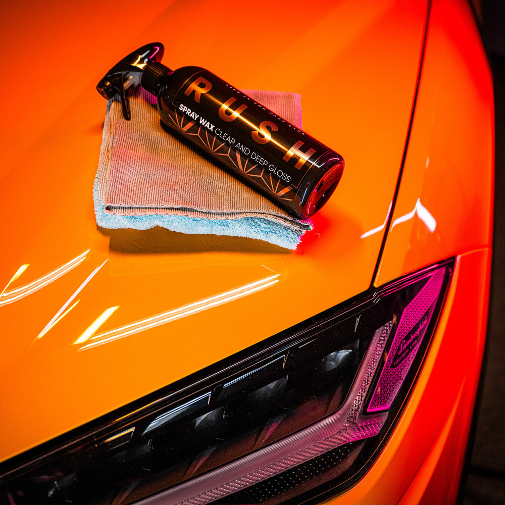 Een fles RUSH Spray Wax en een microvezeldoek liggen op de motorkap, klaar om glanzen te auto laten met een beschermende laag.