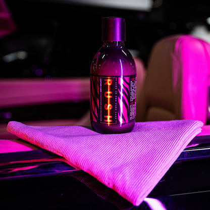 A bottle of the Leather Clean & Care Package rests on a folded cloth atop a car door under vibrant purple-pink lighting, suggesting thorough cleaning for your leather upholstery, with a blurred car interior in the background.