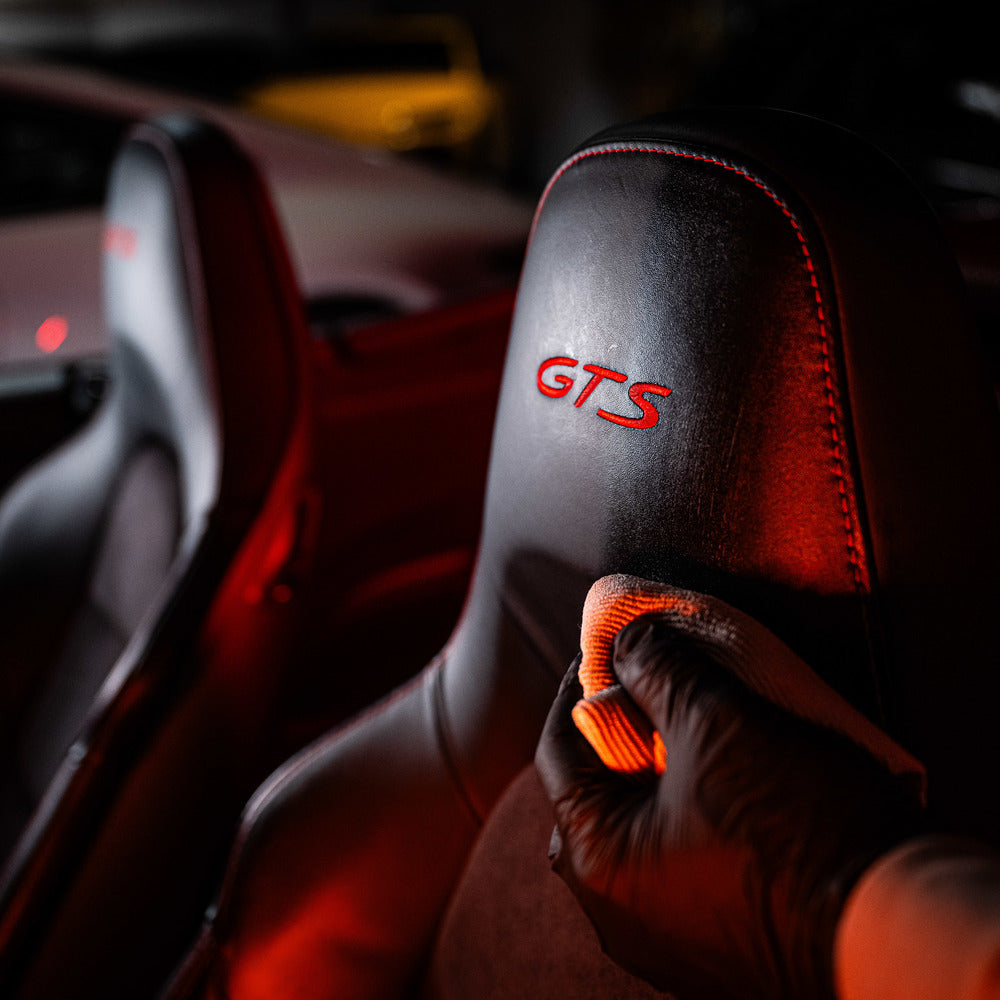 A gloved hand uses the Leather Clean & Care Package to wipe the headrest of a black leather car seat with red GTS embroidery under red lighting, while another car and seat appear in the background.