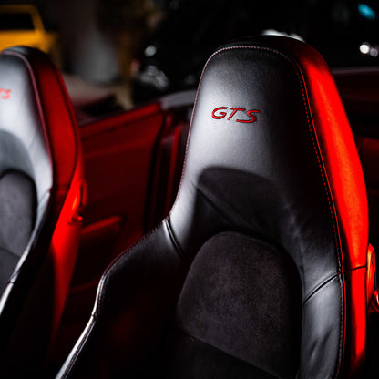 Close-up of black lederen autobekleding with GTS in red on the headrest, lit by red lighting. Another similar seat is partially visible, underlining the importance of goed leer verzorgen with the Leather Clean & Care Package.