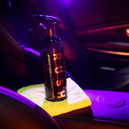 Een spuitfles Rush Interior Clean All Surface Cleaner voor interieur reiniger auto zit op een gele doek op een autostoel onder paarse verlichting.