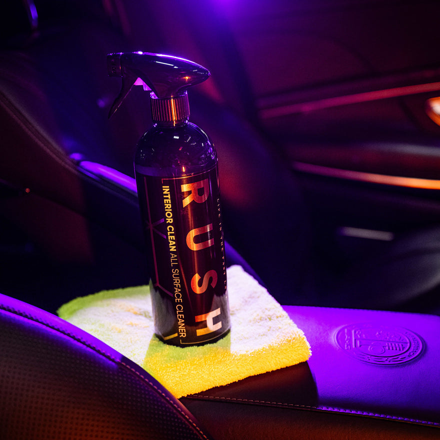 A bottle of Valentijnsdag Package Interior Clean sits on a yellow microfiber cloth atop a car’s dark leather seat under purple and orange lights, ideal for your next auto interieur reinigen or as part of a special gift.
