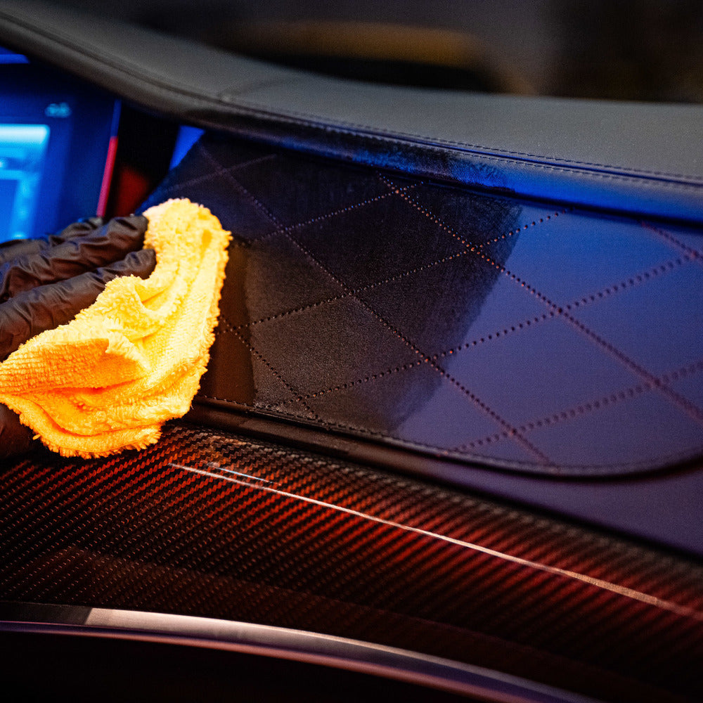 Een gehandschoende hand reinigt het dashboard van een gewatteerde leren auto met een gele microvezeldoek, perfect voor interieurreiniging in de buurt van het touchscreen display.