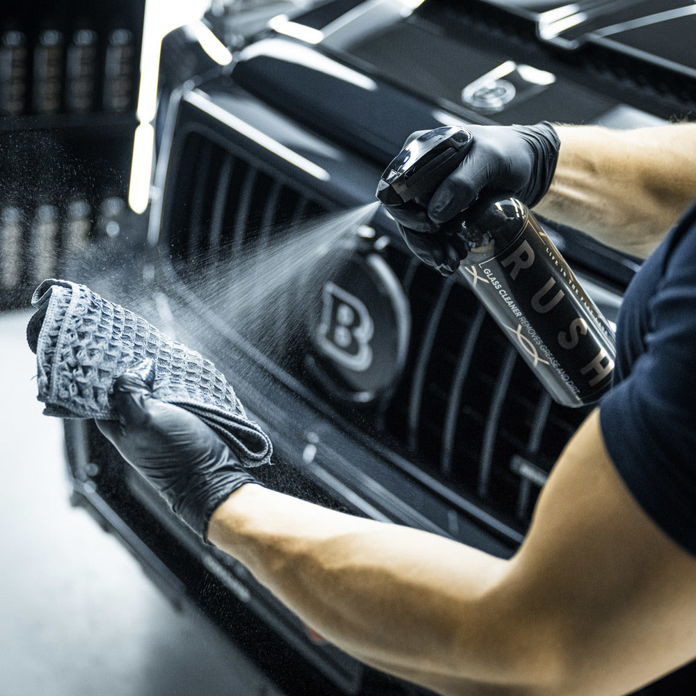 Een persoon met zwarte handschoenen sproeit glasreiniger op een microvezeldoek voor een zwarte grille van een auto.