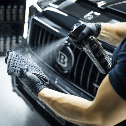 Een persoon met zwarte handschoenen sproeit glasreiniger op een microvezeldoek voor een zwarte grille van een auto.