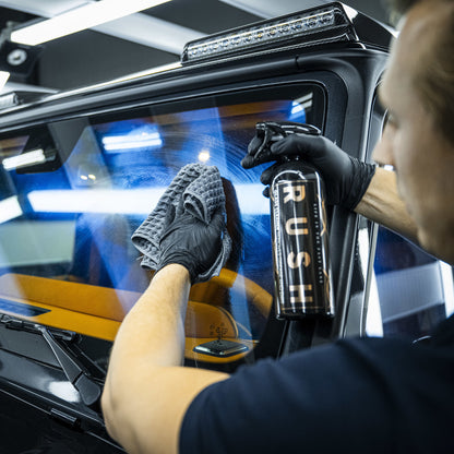 Een persoon met zwarte handschoenen aan reinigt de voorruit van een auto met glasreiniger en een grijs microvezeldoek in een goed verlichte garage.