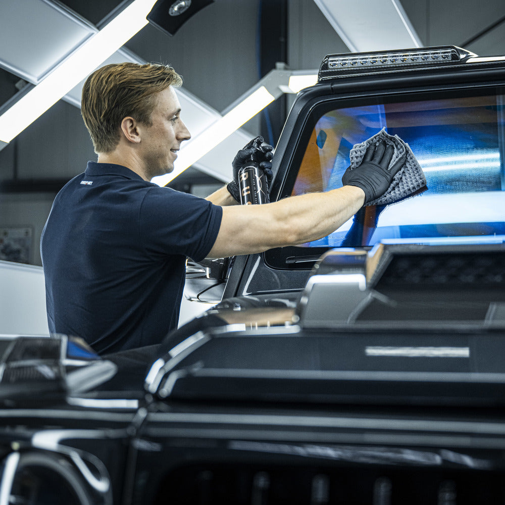 Een man met handschoenen aan maakt een autoruit schoon met een microvezeldoek en glasreiniger in een goed verlichte garage of werkplaats.