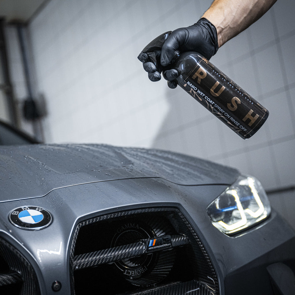 A person wearing a black glove sprays Exterior Package onto the hood of a gray BMW in a tiled garage, ensuring auto reiniging and improved lak bescherming.