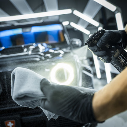 Een persoon met zwarte handschoenen sproeit RUSH Spray Wax op een microvezeldoek, klaar om te auto laten glanzen voor een zwarte auto onder felle lampen.