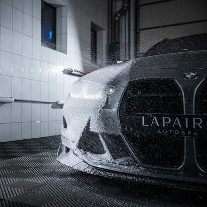 Een BMW-auto wordt gewassen met dik schuim uit een Foam Cannon Pro in een overdekte autowasinstallatie, het sop bedekt de voorkant.