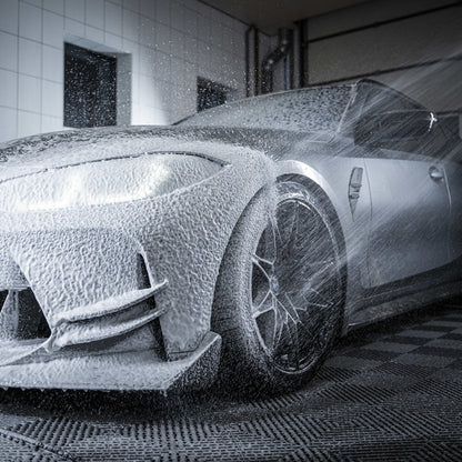 A luxury sports car is coated in thick white foam from the Foam Cannon Package as water sprays over its front wheel and headlights, creating a dynamic, clean atmosphere in an indoor detailing garage.