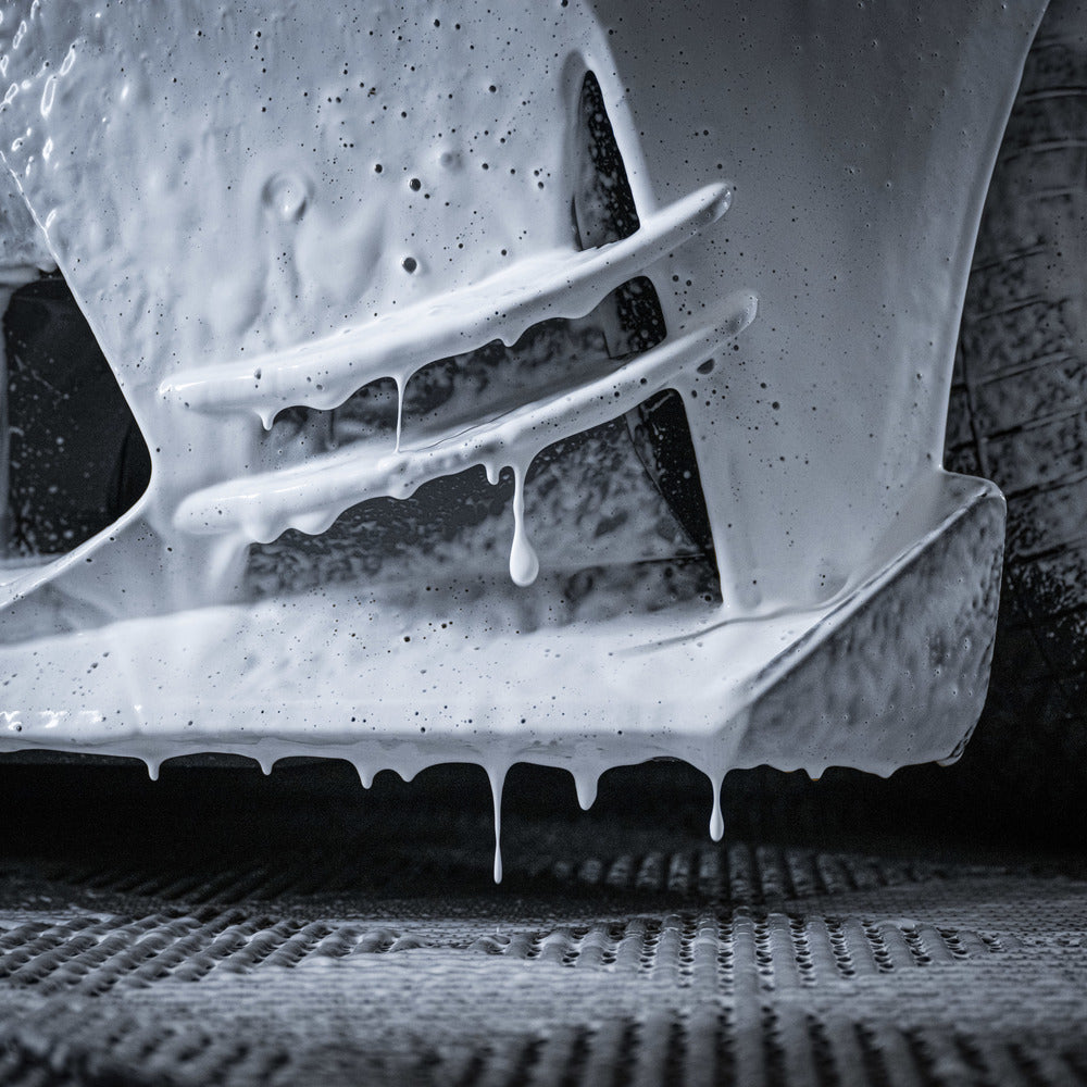 A close-up of a car bumper covered in thick, dripping Snow Foam from the Foam Cannon Package during a pre-wash, with foam streaks and drops visible on the surface.