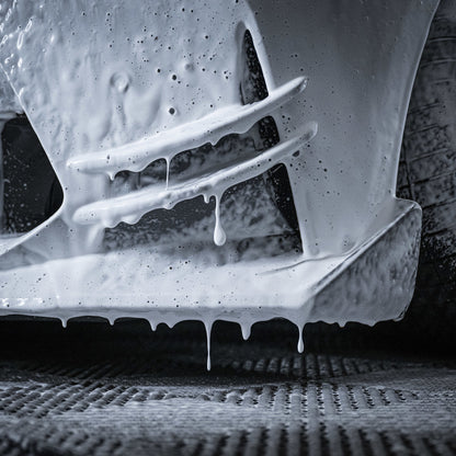 Close-up van een autobumper met dik schuim en water dat eraf druipt tijdens het autowassen, band en natte grond zichtbaar.