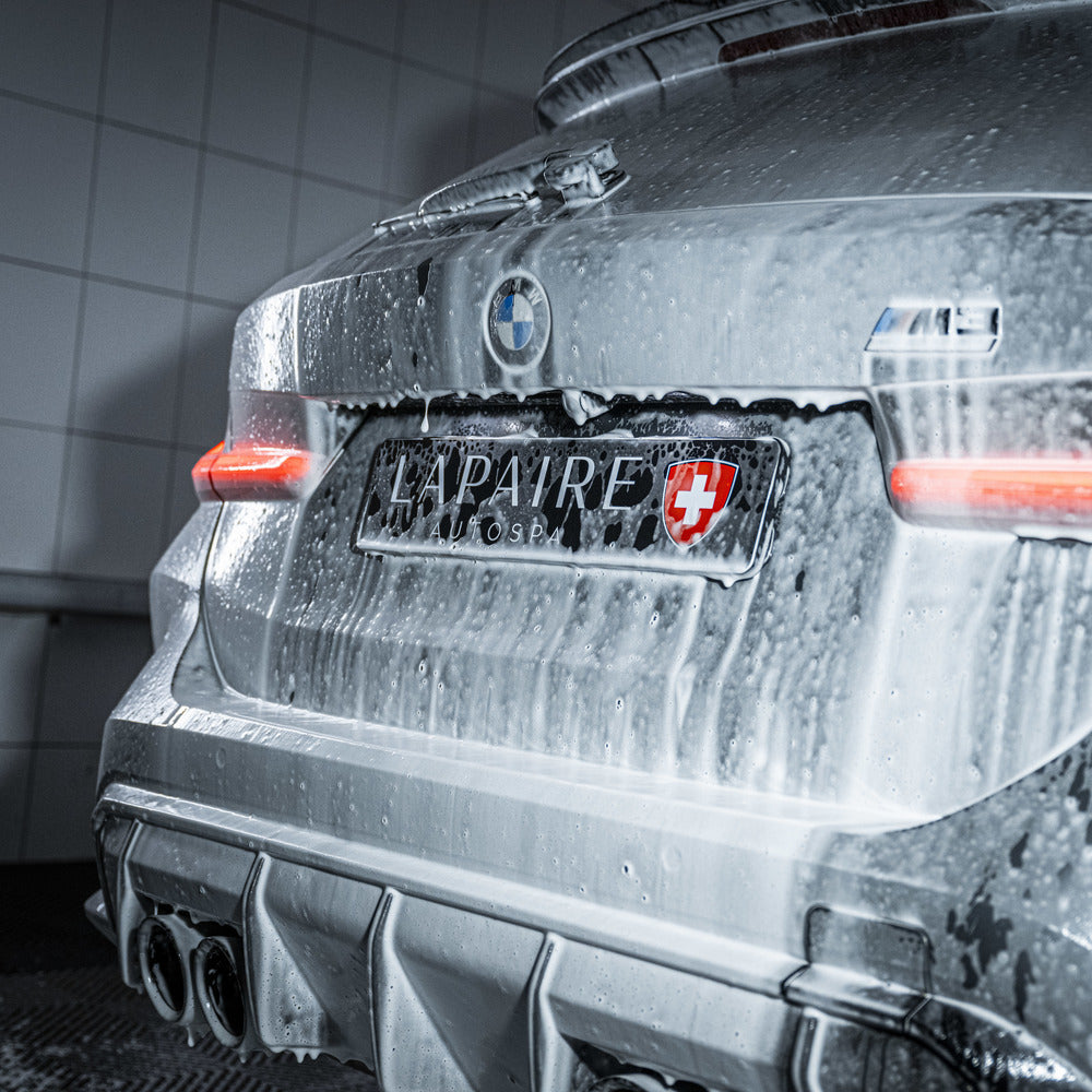 A silver BMW M4, seen from the rear in a car wash with red tail lights on and a Swiss-themed LAPAIRES plate, is covered in thick suds using the Foam Cannon Package, highlighting an auto pre-wash in action.