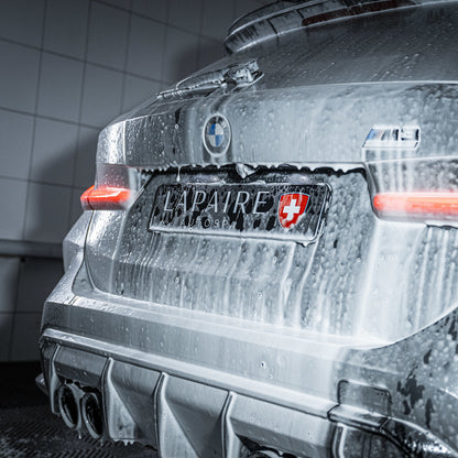 A silver BMW M4, seen from the rear in a car wash with red tail lights on and a Swiss-themed LAPAIRES plate, is covered in thick suds using the Foam Cannon Package, highlighting an auto pre-wash in action.