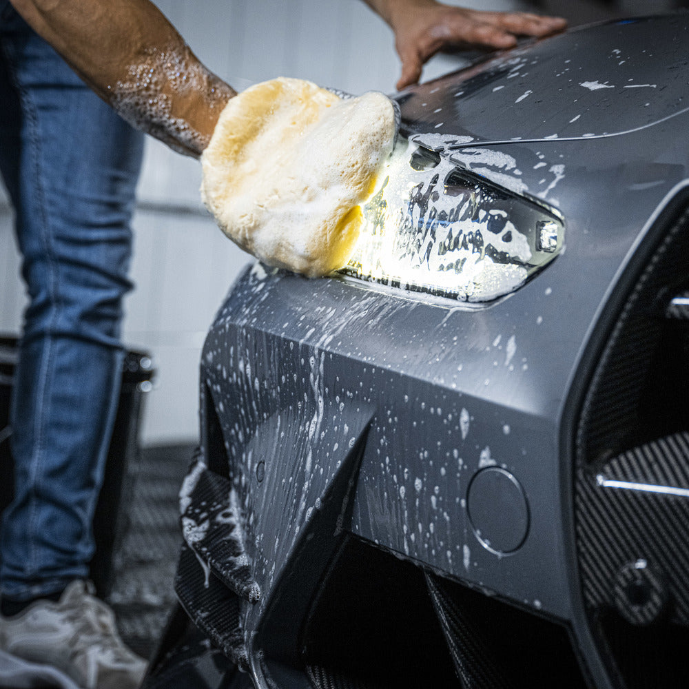 Een persoon gebruikt een wolhandschoen om een zwarte auto te wassen met zeep, waarbij de nadruk ligt op het achterlicht en insectenresten verwijderen.