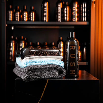 Twee gevouwen handdoeken met Spray Wax en RUSH-flessen op een zwart oppervlak, perfect voor het Showroom Finish-pakket; planken op de achtergrond.
