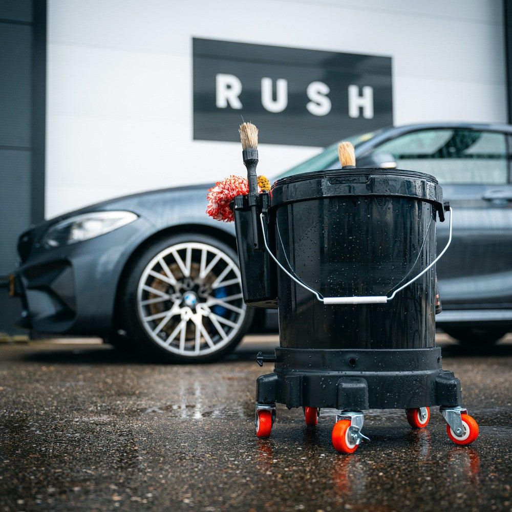 A black Bucket Dolly with brushes and red caster wheels is on wet pavement in front of a gray car and a building with a RUSH sign.