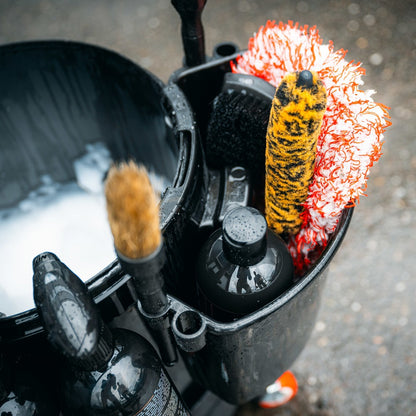 Een zwarte emmer met flessen, borstels en een geel-witte washandschoenhouder staat gevuld met zeepsop op een natte stoep.