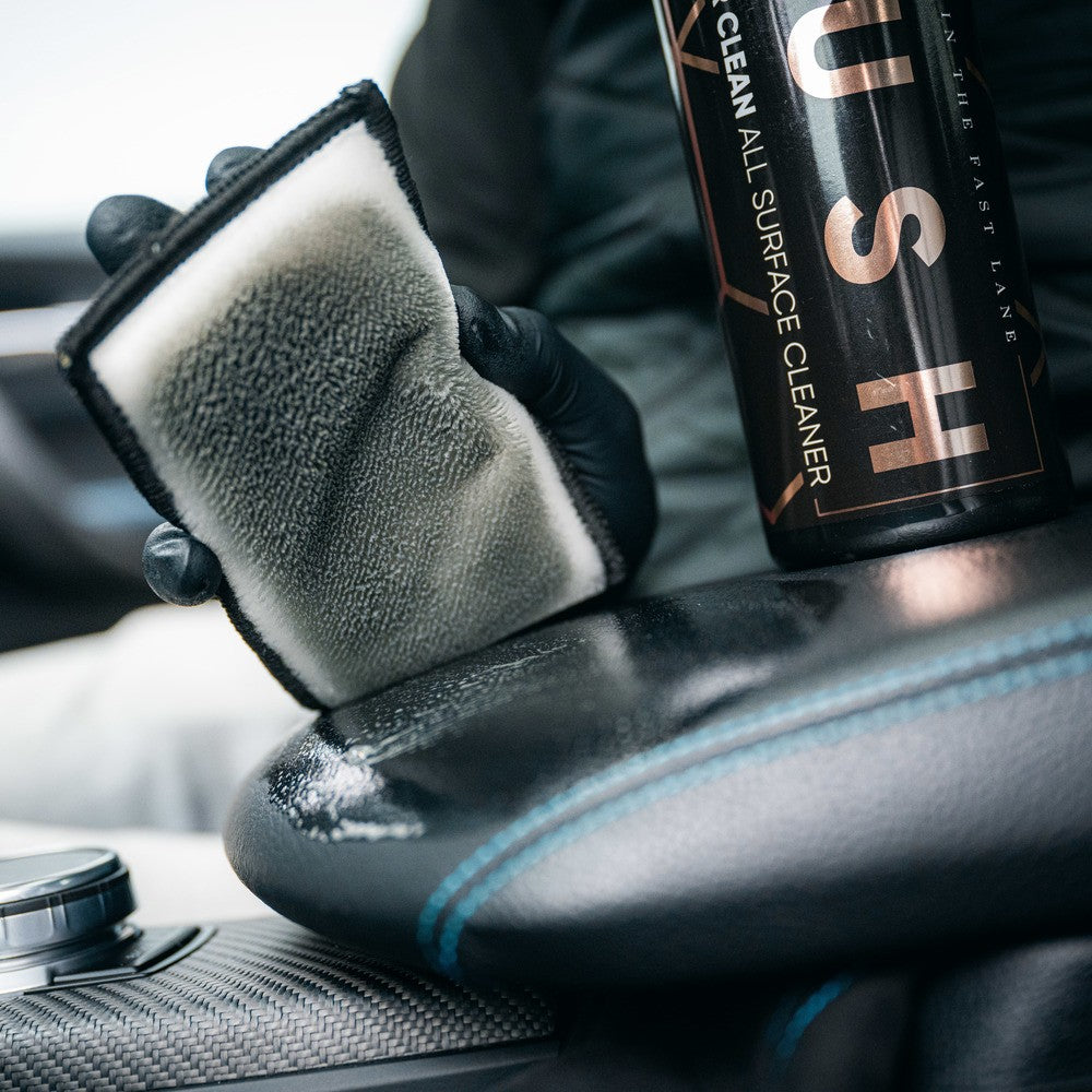 A gloved hand holds a dirty 2-in-1 Interior Scrubbing Pad - 2-pack above a car’s leather seat, next to a bottle labeled CLEAN ALL SURFACE CLEANER, illustrating the interior cleaning process.