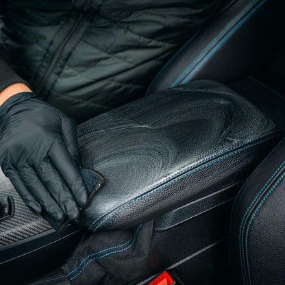 Wearing black gloves, a person cleans a black leather car armrest with blue stitching using the 2-in-1 Interior Scrubbing Pad - 2-pack for effective interior auto care.