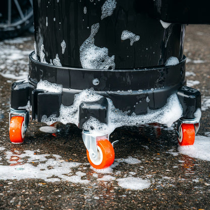 Close-up of a black cleaning bucket on a Bucket Dolly with rode zwenkwielen, covered in soapy suds and water, resting on a wet, textured concrete surface.