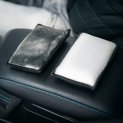 Two rectangular 2-in-1 Interior Scrubbing Pads (2-pack) are on a car’s black leather armrest—one dirty gray, one clean white. Clothing and a seat in the background highlight the pad’s cleaning effect.