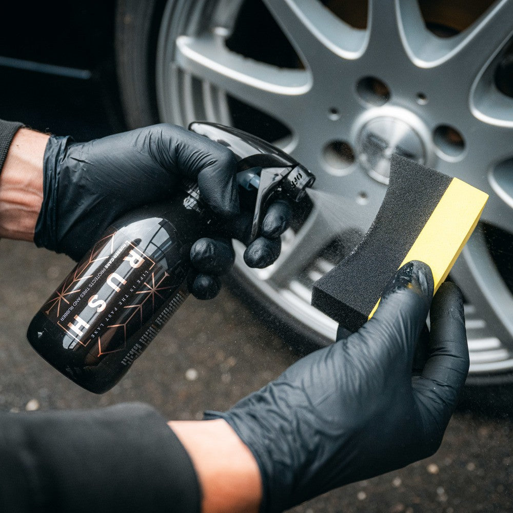 Een persoon met zwarte handschoenen gebruikt een rubberen onderhoudsspuit om reinigingsmiddel op een spons te spuiten bij een autowiel met een zilveren rand.