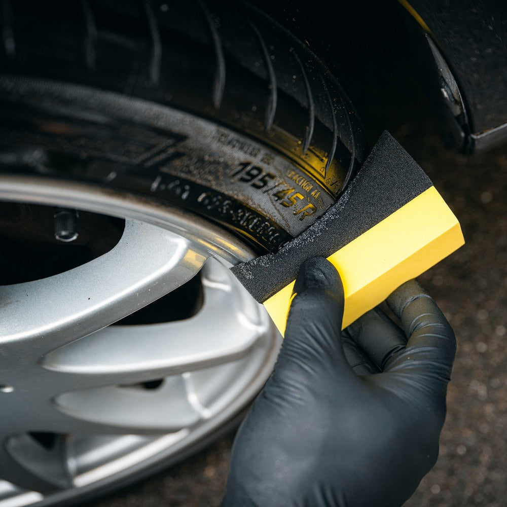 Een gehandschoende hand gebruikt een bandenzwart applicator om product te reinigen of aan te brengen op een autoband en een zilverkleurige lichtmetalen velg.