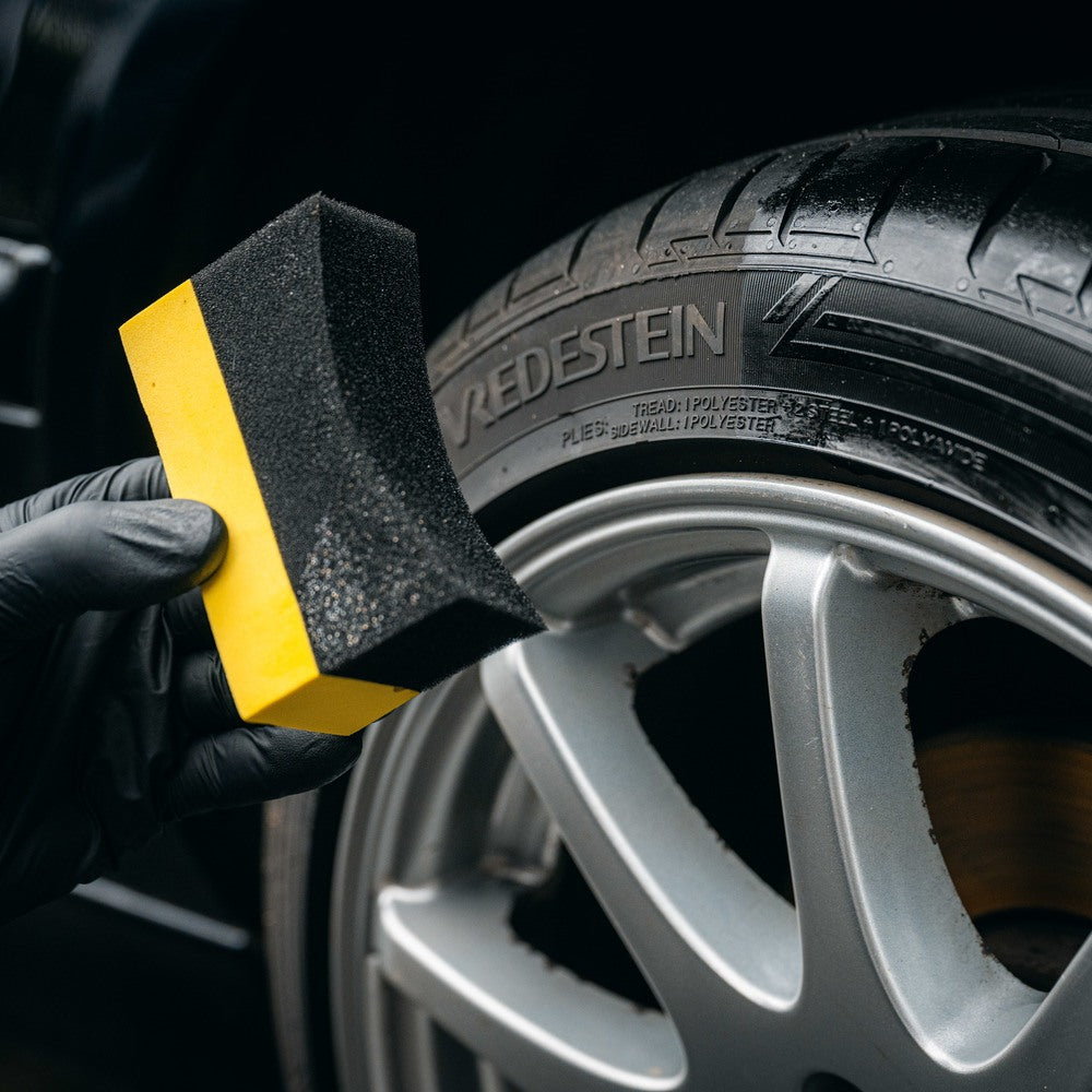 Een gehandschoende hand houdt een bandenzwart-applicator bij een autoband en een zilverkleurige lichtmetalen velg voor reiniging of detailing.