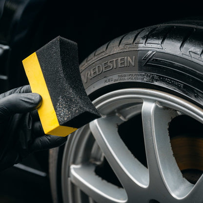 Een gehandschoende hand houdt een bandenzwart-applicator bij een autoband en een zilverkleurige lichtmetalen velg voor reiniging of detailing.