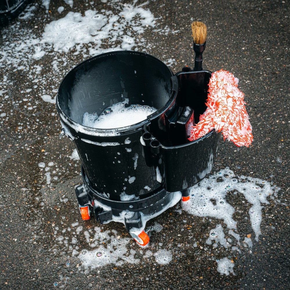 Een zwarte opbergbak emmer met zeepsop, een schoonmaakdoekje en een borstel zit op een natte stoep omringd door zeepsop.
