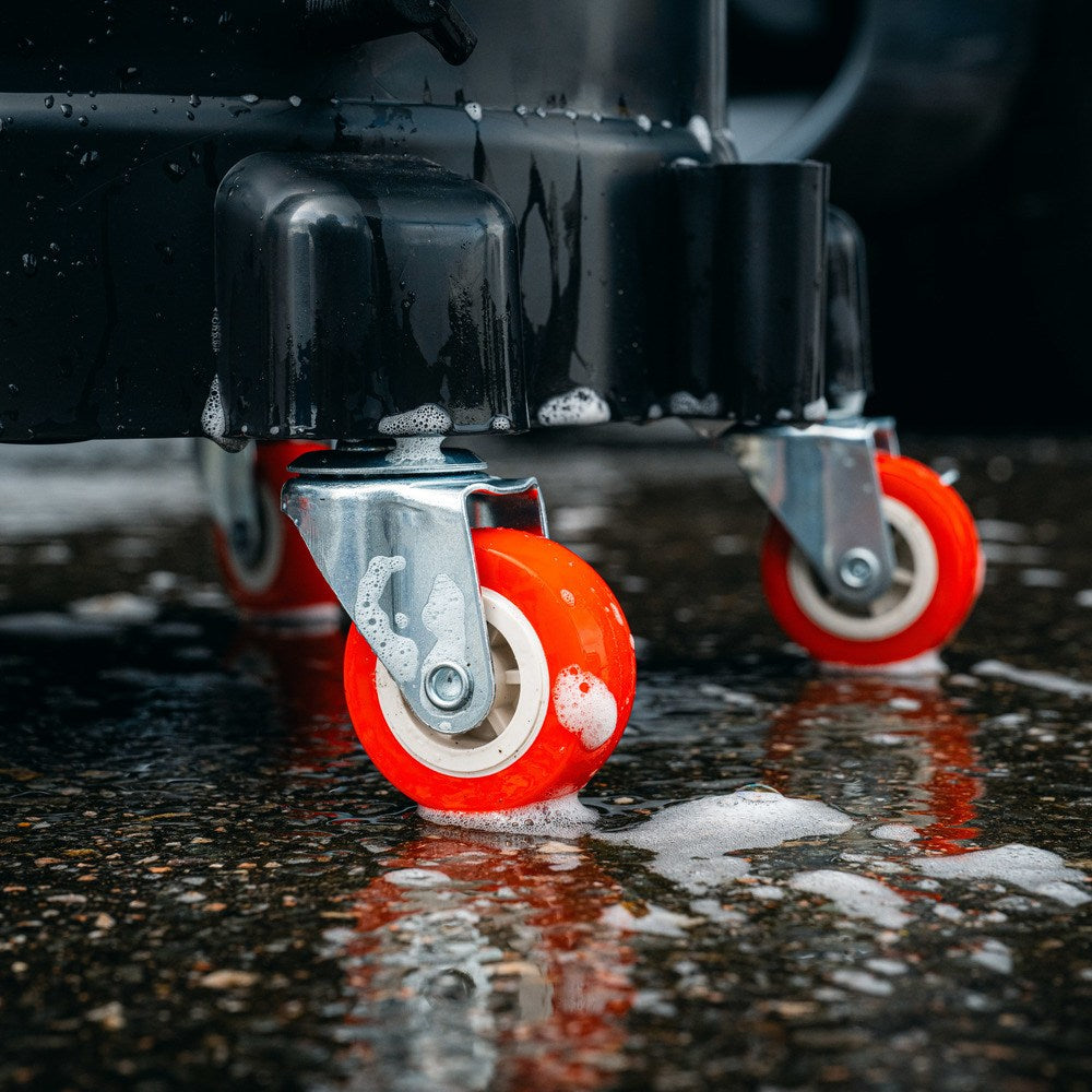 Close-up of two bright red swiveling wheels on a black Bucket Dolly, positioned on a wet, soapy floor with bubbles and reflections—ideal for easily moving your cleaning bucket.
