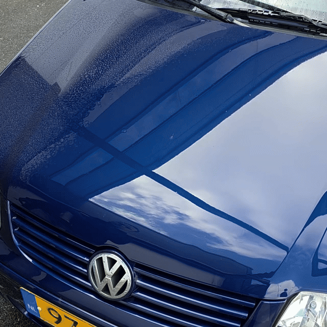 Close-up of the hood of a blue Volkswagen with water droplets—thanks to the Black Friday Package—plus building and sky reflections on the glossy surface. Part of the license plate and headlight are visible.