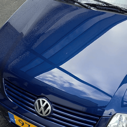 Close-up of the hood of a blue Volkswagen with water droplets—thanks to the Black Friday Package—plus building and sky reflections on the glossy surface. Part of the license plate and headlight are visible.